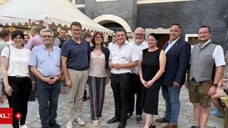 Eine Gruppe von formell gekleideten Menschen posiert für ein Foto vor einem Gebäude mit Zelten und Personen im Hintergrund.