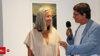 Ein Mann mit einem Mikrofon interviewt eine Frau mit grauen Haaren. Sie stehen vor einer weißen Wand mit einem Bilderrahmen.