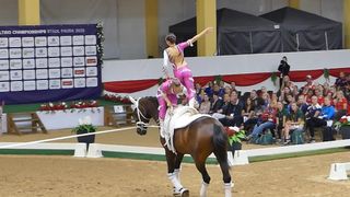 Zwei Frauen in pinkfarbenen Kostümen führen eine Balance-Aktion auf einem braunen Pferd bei einer Veranstaltung durch. Sie stehen vor einem Publikum, das auf Bänken sitzt.