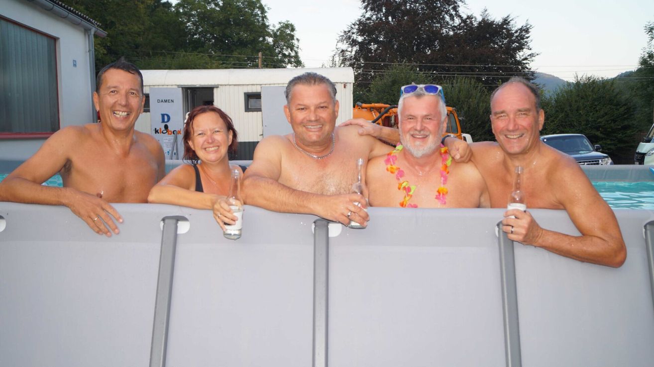Drei Männer und eine Frau lächeln und posieren in einem tragbaren Pool. Die Männer sind ohne Hemd, einer trägt eine Halskette und ein anderer eine Girlande. Die Frau hält ein Glas. Sie sind von Bäumen und einem Gebäude mit einem Schild, das 'DAMEN' steht, umgeben.