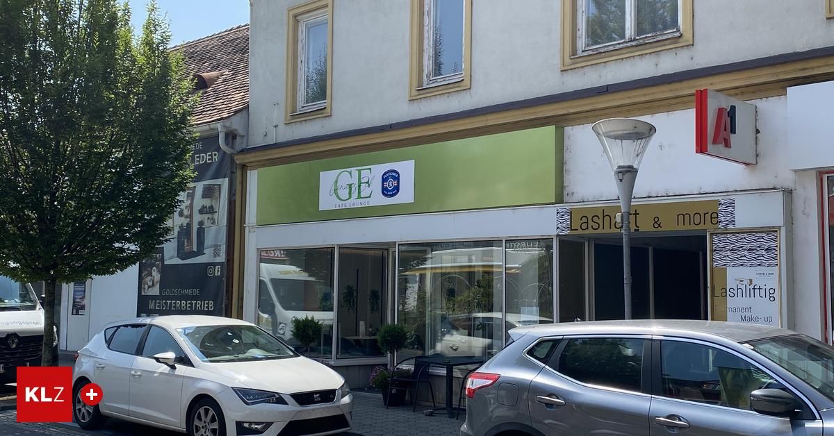 Ein Laden mit 'Age Cafe Lounge'-Schild, grün und weiß, geparkte Autos, Straßenlaterne, Bäume und Pflanzen.