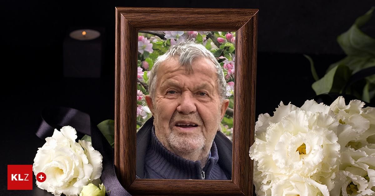 Ein Porträt eines älteren Mannes mit grauem Haar und Bart, der einen blauen Pullover trägt, in einem Holzrahmen mit rosa Blumen und einer Kerze im Hintergrund.