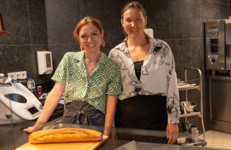 Zwei Frauen in einer Küche, lächelnd und neben einem Schneidebrett mit einem Laib Brot stehend.