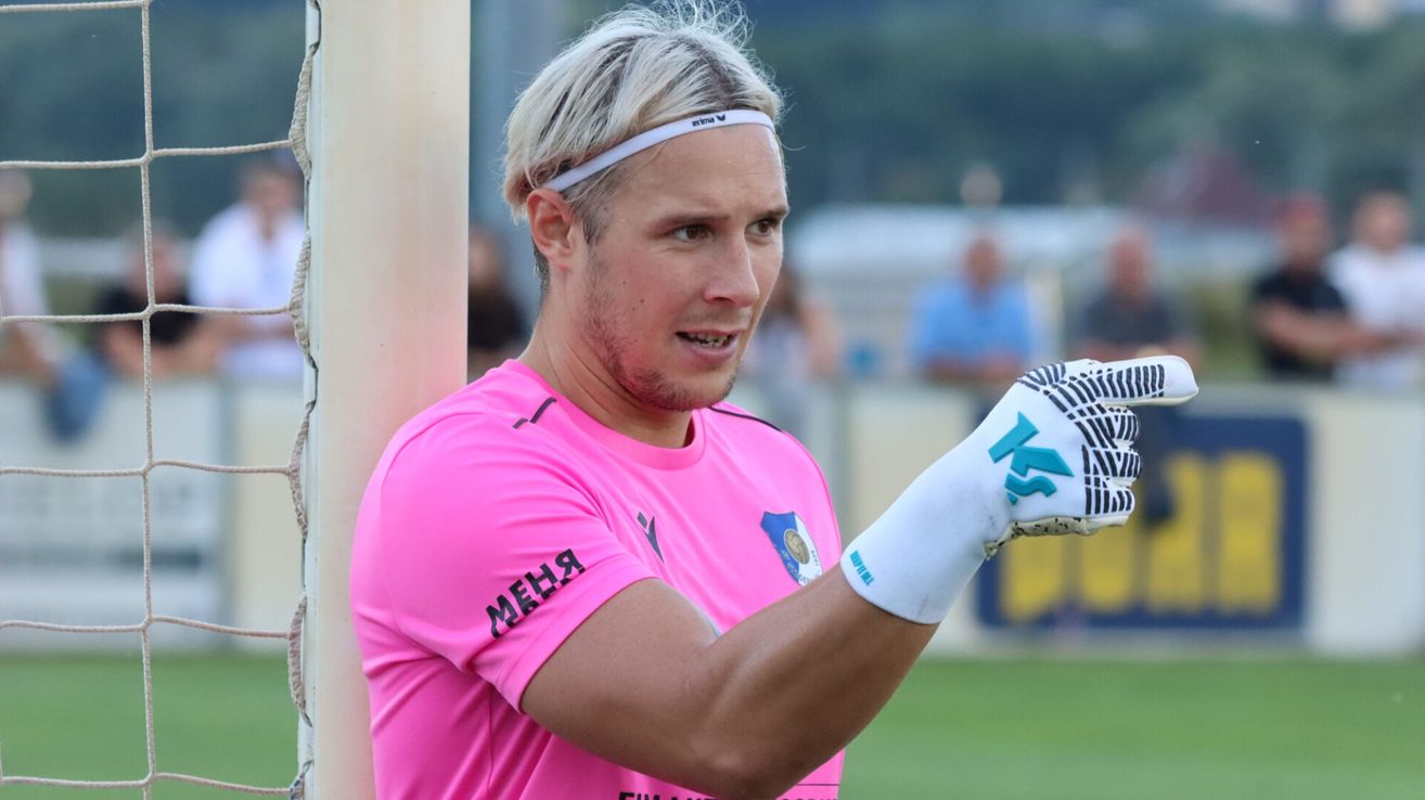 Ein Fußballspieler in einem pinken Trikot und weißen Handschuhen steht und blickt nach vorne, mit einem verschwommenen Hintergrund von Menschen und einem Zaun.