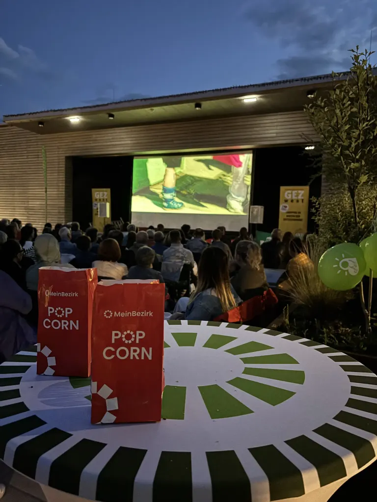 Eine Open-Air-Filmvorführung mit einem Projektor, der einen Fuß zeigt. Das Publikum sitzt, und zwei rote Popcorntüten stehen auf einem Tisch. Es gibt grüne Luftballons in der Nähe.