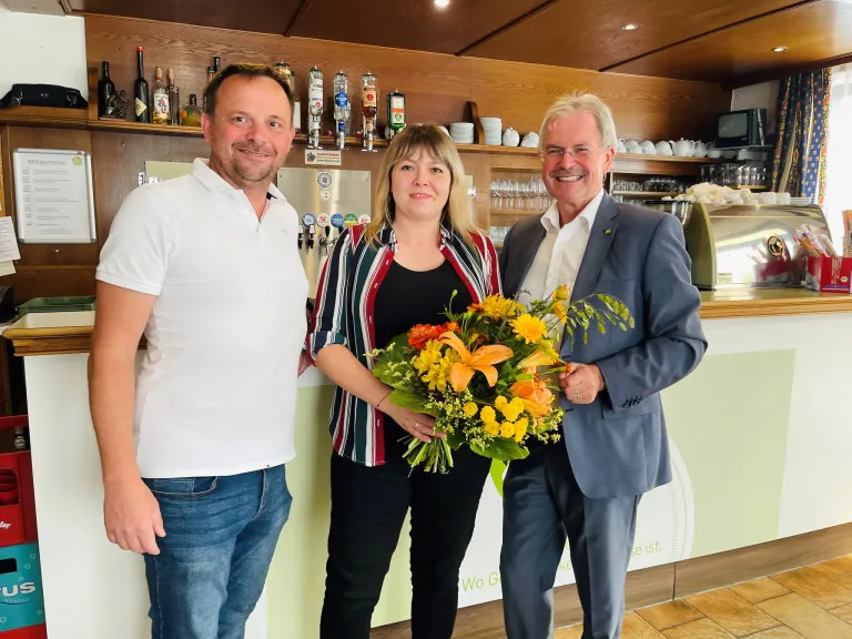 Drei Personen, zwei Männer und eine Frau, stehen gemeinsam in einem Restaurant. Die Frau hält einen Strauß gelber und orangefarbener Blumen. Hinter ihnen befindet sich eine Theke mit Regalen, die mit Flaschen und Tassen bestückt sind.