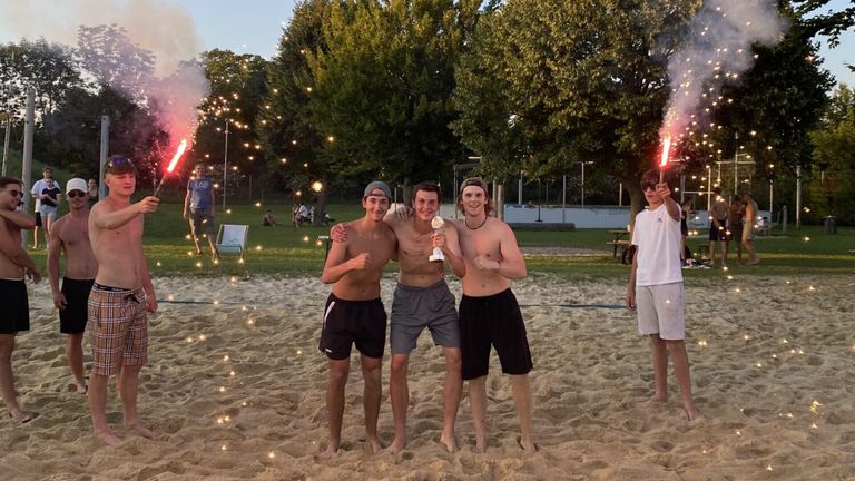 Drei junge Männer stehen im Sand, halten einen Pokal und posieren für ein Foto. Dahinter feiert eine Menge mit Feuerwerk.