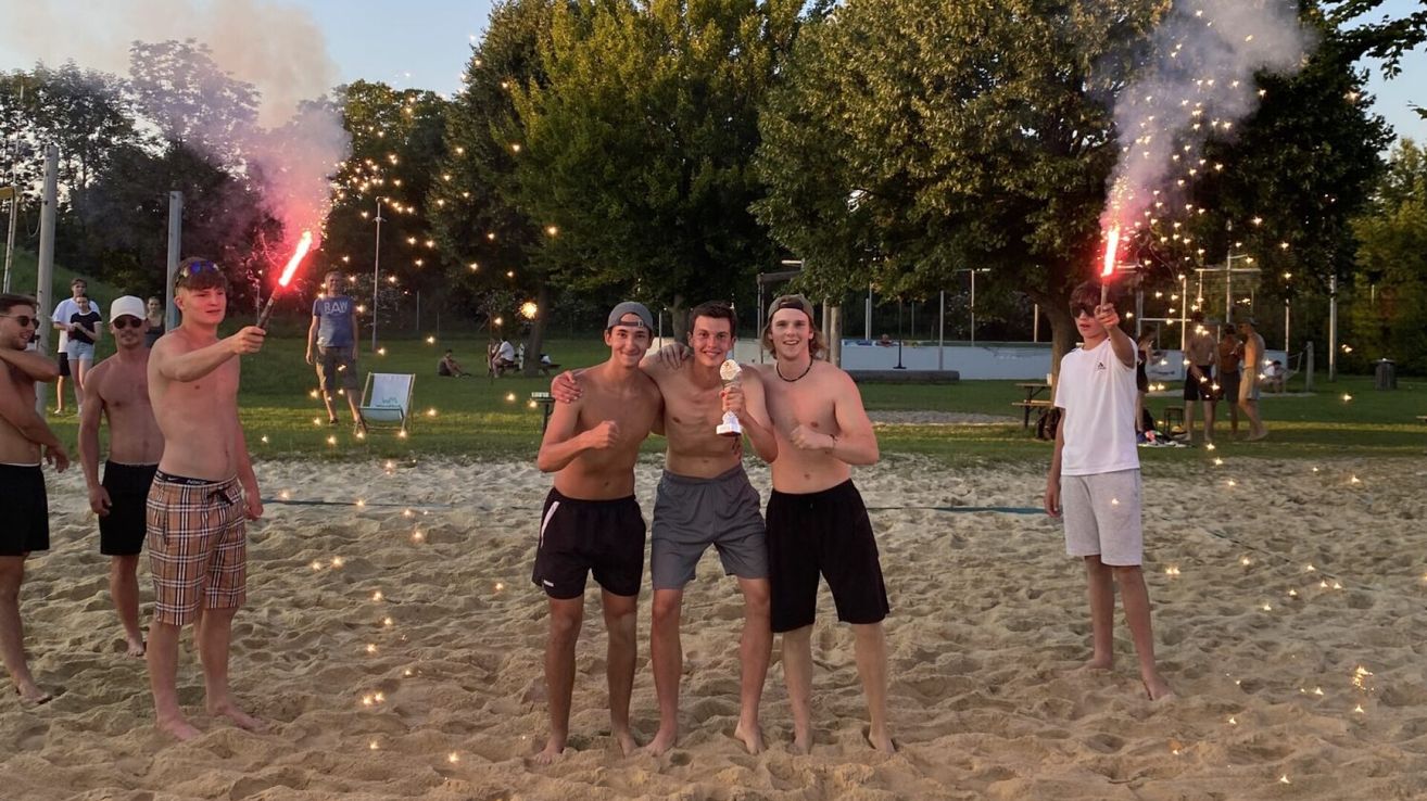 Drei junge Männer stehen im Sand, halten einen Pokal und posieren für ein Foto. Dahinter feiert eine Menge mit Feuerwerk.