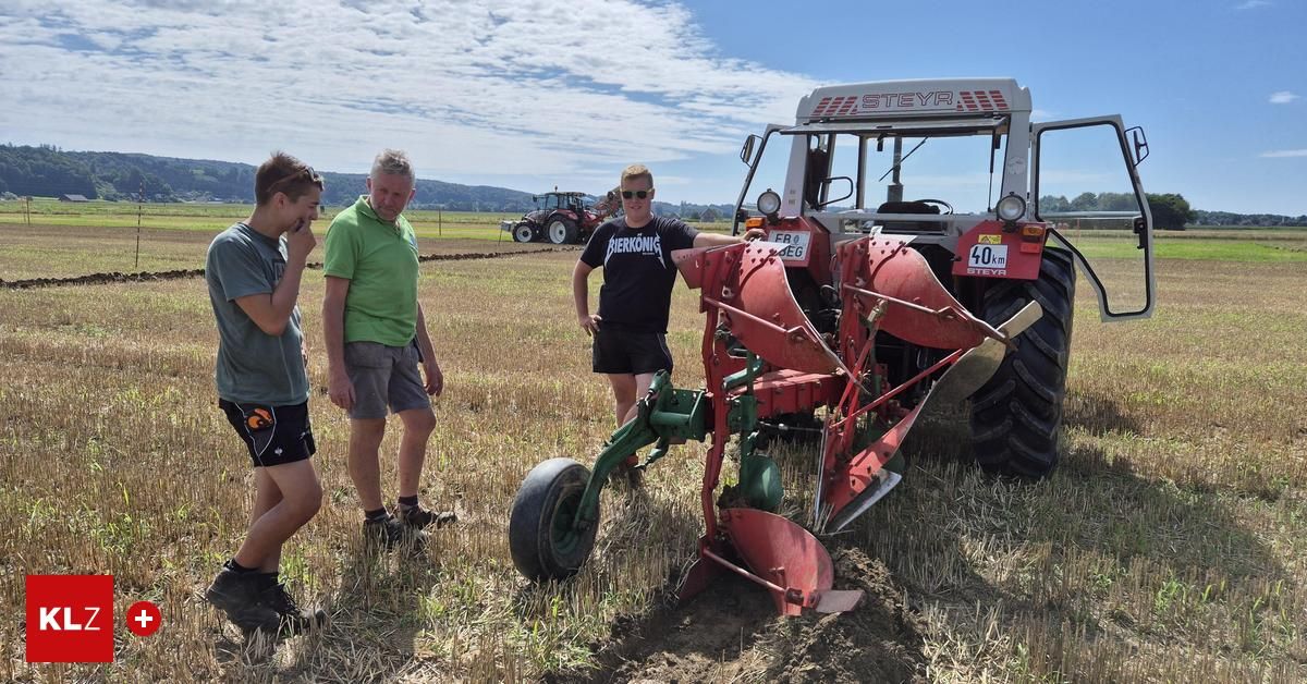 Drei Männer stehen auf einem Feld, einer hält einen roten Pflug. Der Mann in der Mitte trägt ein schwarzes Shirt mit einem weißen Logo. Hinter ihnen steht ein Traktor mit dem Wort STEYR an seiner Seite.