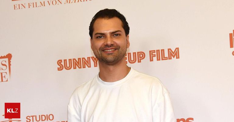 Ein lächelnder Mann in einem weißen T-Shirt steht vor einem weißen Hintergrund mit orangefarbenem Text, der 'SUNNY UP FILM' lautet.