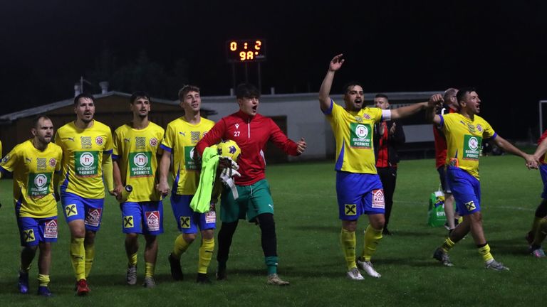 Eine Fußballmannschaft feiert auf einem Feld bei Nacht. Sie tragen gelbe und blaue Trikots mit der Nummer 27 in Rot. Eine Anzeigetafel zeigt 0:2.