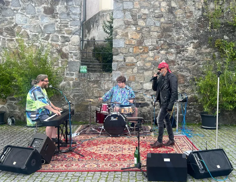 Eine Band tritt auf einem roten Teppich im Freien auf. Ein Schlagzeuger und ein Keyboarder spielen links, während ein Sänger mit einem Mikrofon rechts steht. Eine Steinmauer und eine Treppe sind im Hintergrund zu sehen.