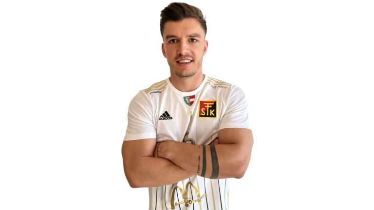 Ein Mann in einem weißen Adidas-Fußballtrikot mit gelbem Logo, Arme verschränkt, lächelt.