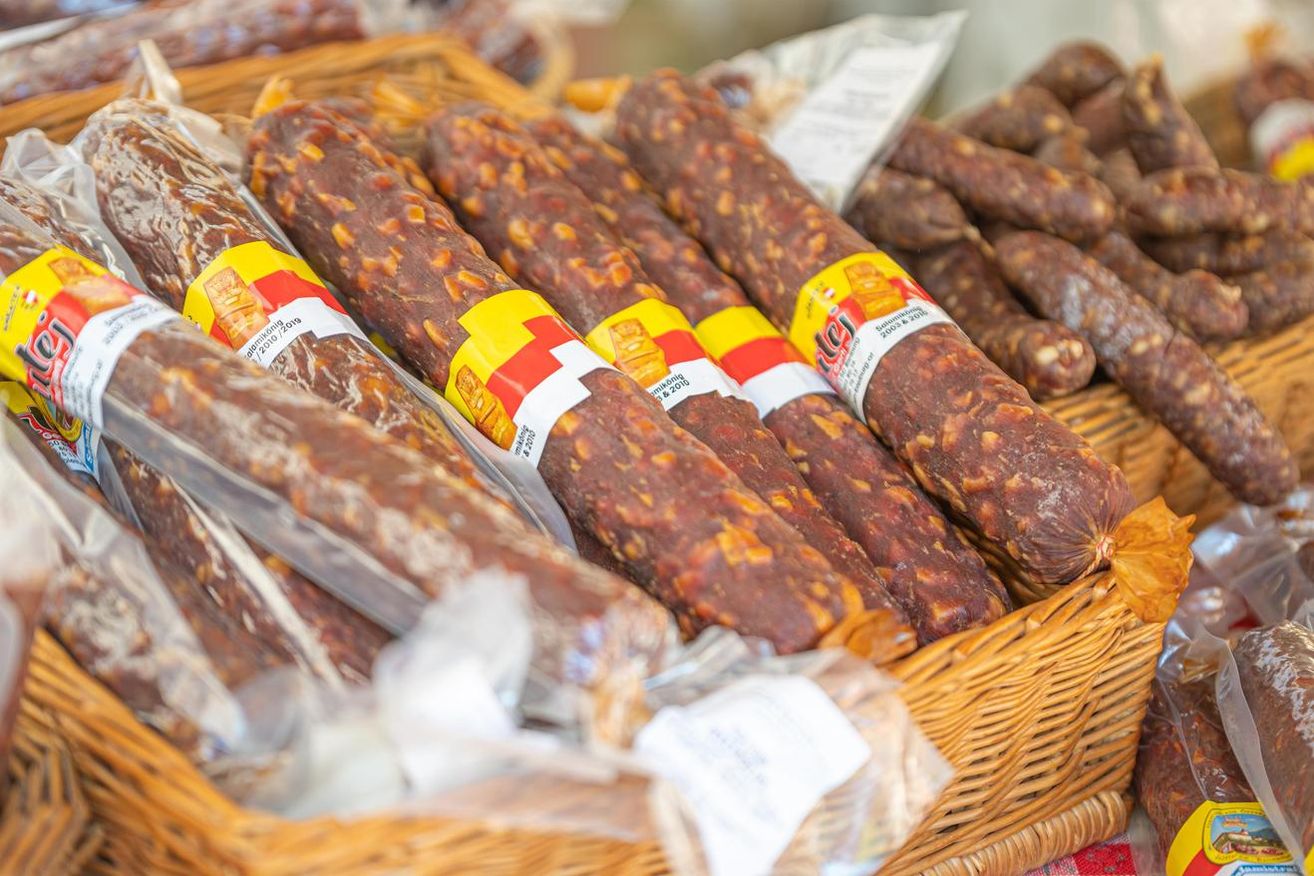 Ein Nahaufnahme-Blick auf mehrere lange, zylindrische Würste, die in Plastik eingewickelt und in einem Weidenkorb angeordnet sind. Jeder Wurst hat ein Etikett mit roten, gelben und weißen Farben.