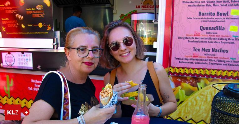 Zwei Frauen sitzen an einem Tisch in einem Imbisswagen, eine lächelt und hält ein Sandwich, die andere trägt eine Brille. Ein Speisezettel ist hinter ihnen, und eine Flasche steht auf dem Tisch.