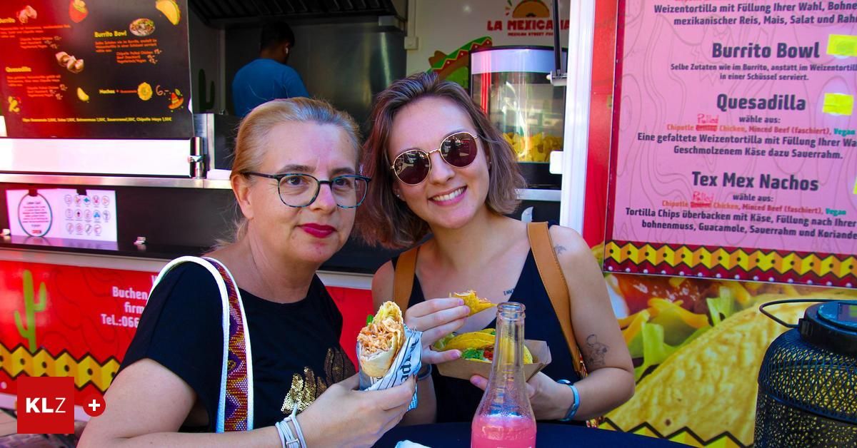 Zwei Frauen sitzen an einem Tisch in einem Imbisswagen, eine lächelt und hält ein Sandwich, die andere trägt eine Brille. Ein Speisezettel ist hinter ihnen, und eine Flasche steht auf dem Tisch.