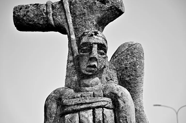 Ein Schwarz-Weiß-Foto zeigt eine verwitterte Statue eines Engels mit Flügeln, der eine Lanze hält, an einem Kreuz befestigt.