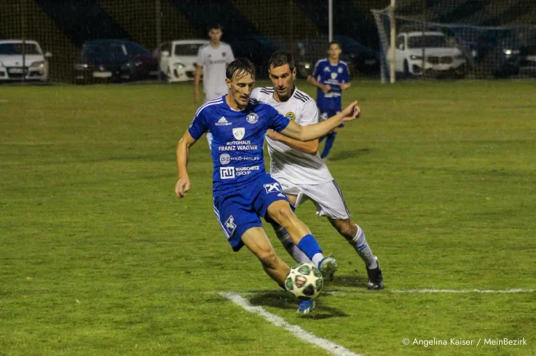 Zwei Fußballspieler spielen auf einem Feld im Regen. Der Spieler in Blau versucht, den Ball zu schießen.