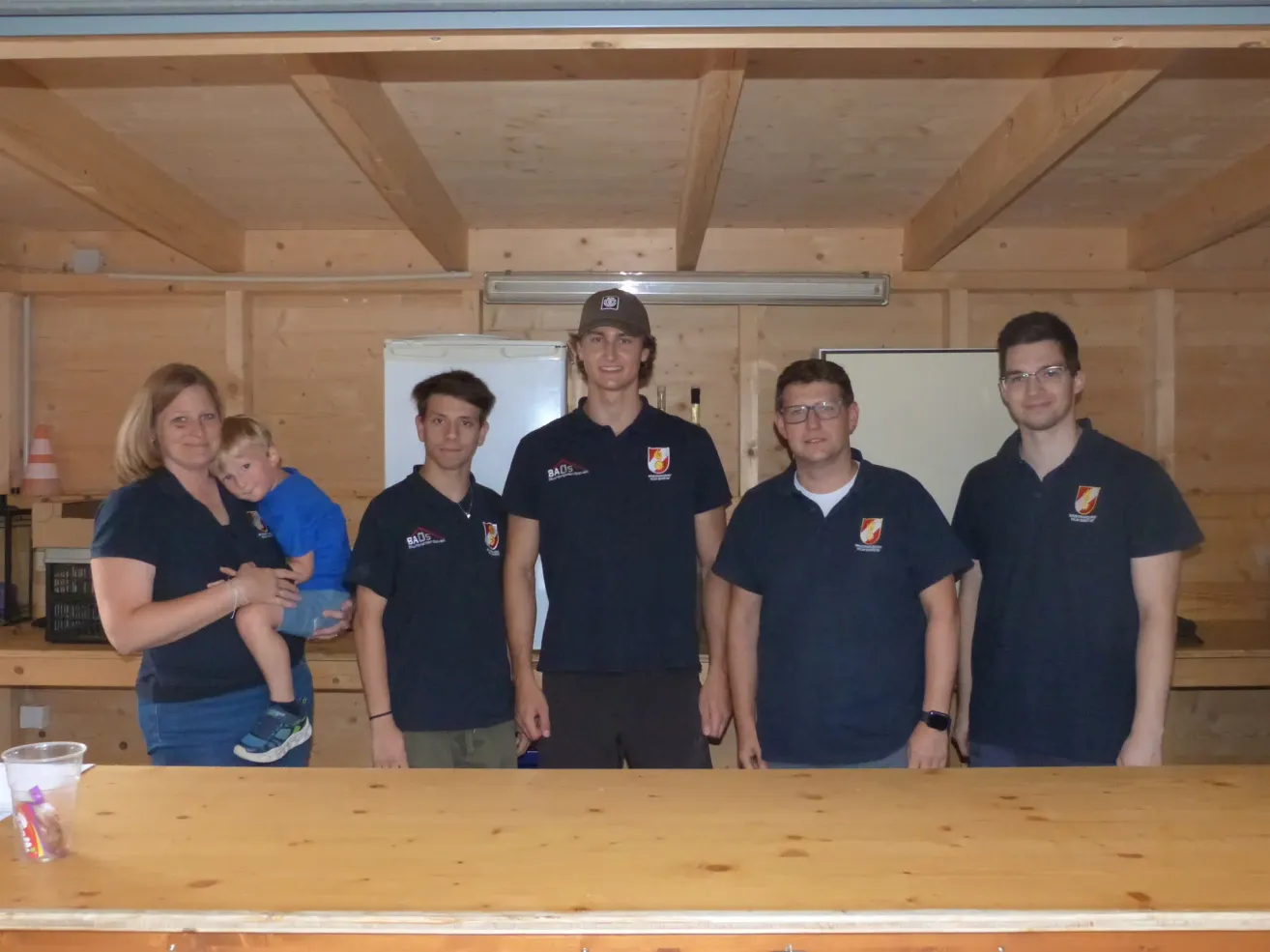 Eine Gruppe von Personen in identischen blauen Shirts mit weißen Logos posiert für ein Foto in einem Raum mit Holzwänden und einer Decke.