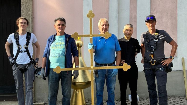 Vier Männer stehen vor einer Kirche und halten ein goldenes Kreuz. Zwei tragen Handschuhe und zwei tragen Freizeitkleidung. Hinter ihnen steht eine goldene Kuppel.