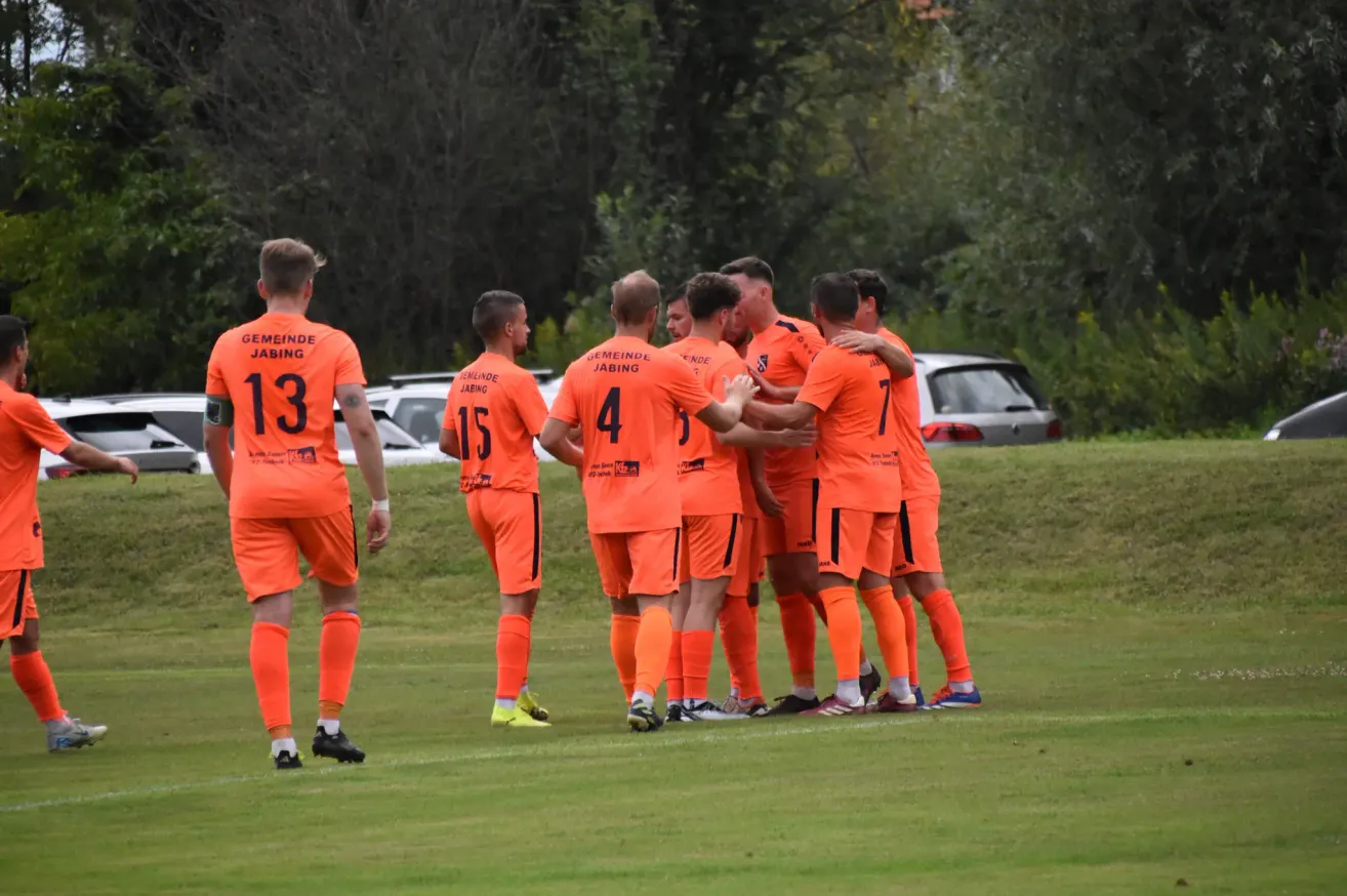 Eine Gruppe von Fußballspielern in orangefarbenen Trikots steht auf einem Rasen. Einer von ihnen trägt ein Trikot mit der Nummer 13, und andere tragen die Nummern 4, 7 und 15. Sie scheinen zu feiern. Dahinter sind zwei Autos geparkt.