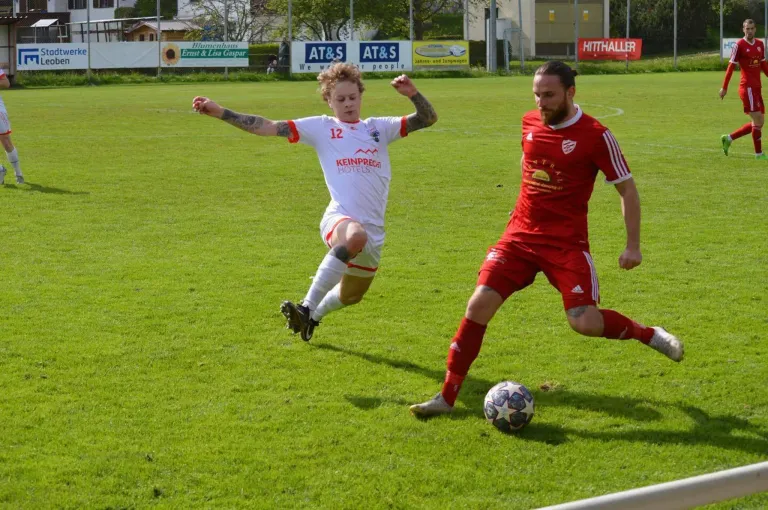 Zwei Fußballspieler auf einem Feld. Einer in Weiß mit 'Keinprege Hotels' auf seinem Trikot, der andere in Rot, beide laufen auf einen Fußball zu.