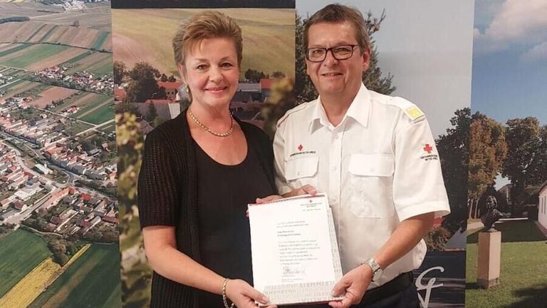 Eine Frau mit kurzen Haaren und einer Halskette hält ein Zertifikat mit einem Mann in einer weißen Uniform mit einem roten Kreuz auf der Schulter.