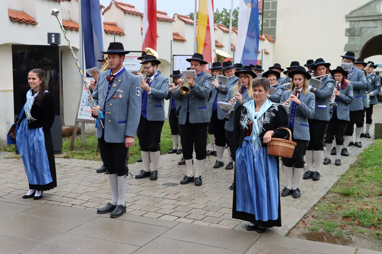 Eine Blaskapelle in traditioneller bayerischer Tracht spielt auf einem gepflasterten Bereich. Die Band besteht aus Blech- und Holzblasinstrumenten. Die Mitglieder tragen Uniformen mit Hüten und Schuhen. Eine Frau steht vor ihnen mit einem Korb.