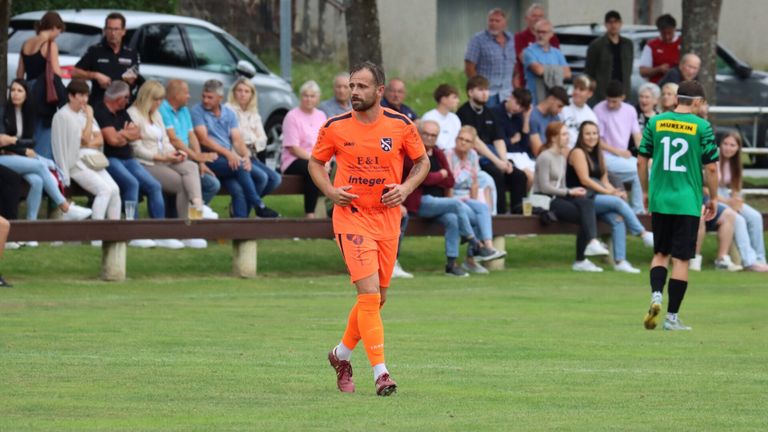 Ein Fußballspieler in orangefarbener Uniform läuft auf dem Spielfeld. Zuschauer sitzen auf den Bänken hinter ihm und beobachten das Spiel.