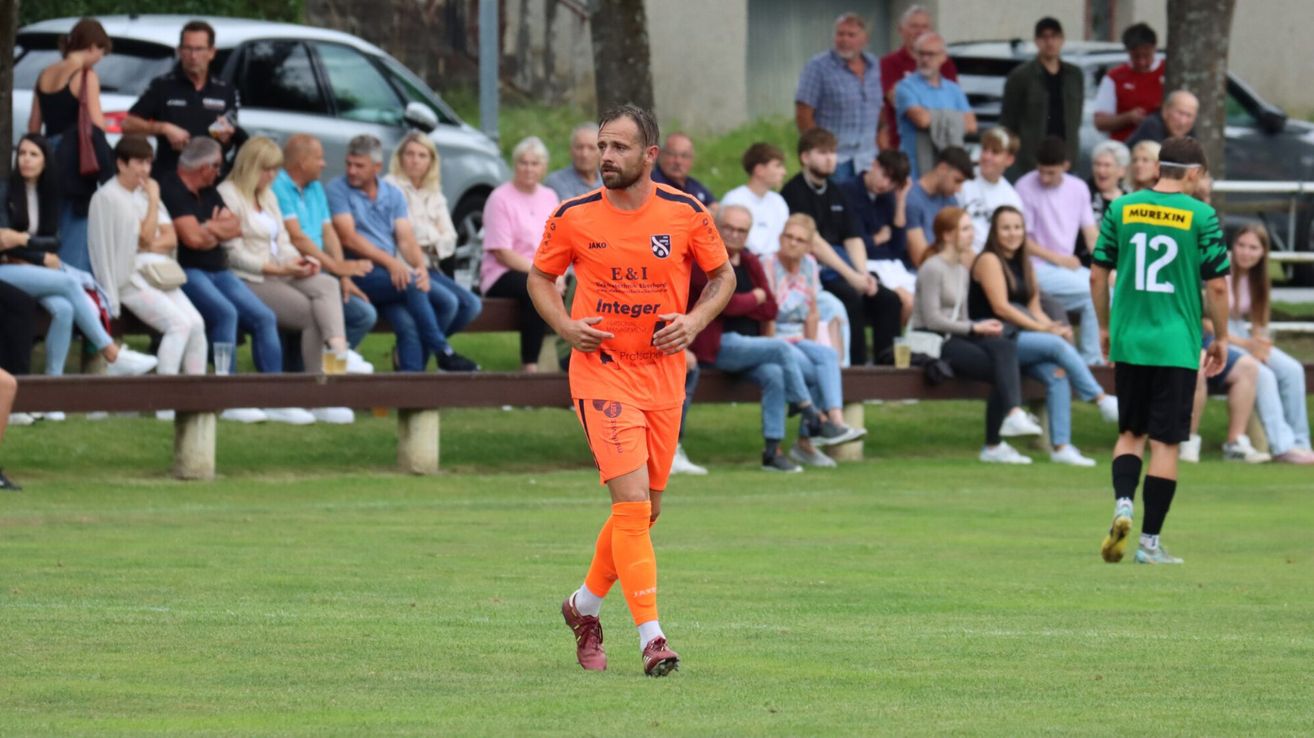 Ein Fußballspieler in orangefarbener Uniform läuft auf dem Spielfeld. Zuschauer sitzen auf den Bänken hinter ihm und beobachten das Spiel.