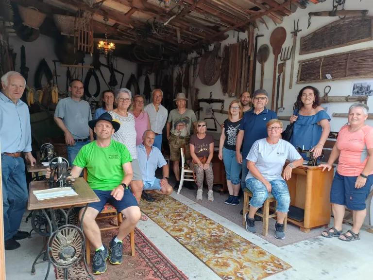 Eine Gruppe von Menschen posiert für ein Foto in einer Werkstatt. Sie stehen und sitzen in einem Raum mit alten Werkzeugen, die an der Wand hängen.