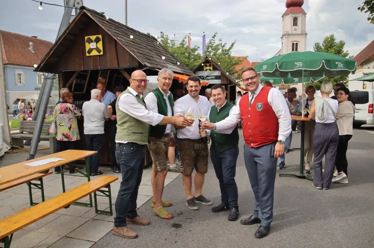 Vier Männer in traditioneller bayerischer Kleidung stoßen mit Wein an. Sie stehen vor einem Holzzelt mit einem gelben Schild. Andere Menschen und eine Kirche sind im Hintergrund zu sehen.
