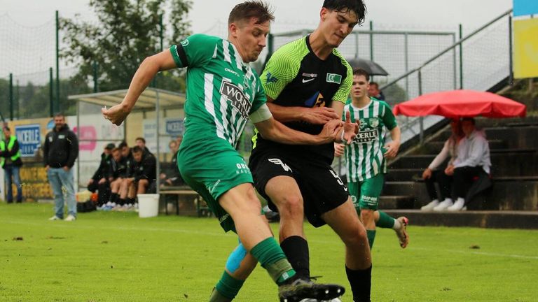 Zwei Fußballspieler sind in einem Spiel engagiert, einer trägt ein grünes Trikot und der andere ein schwarz-grünes Trikot. Beide sind in Aktion, einer versucht den Ball zu schießen, während der andere verteidigt.