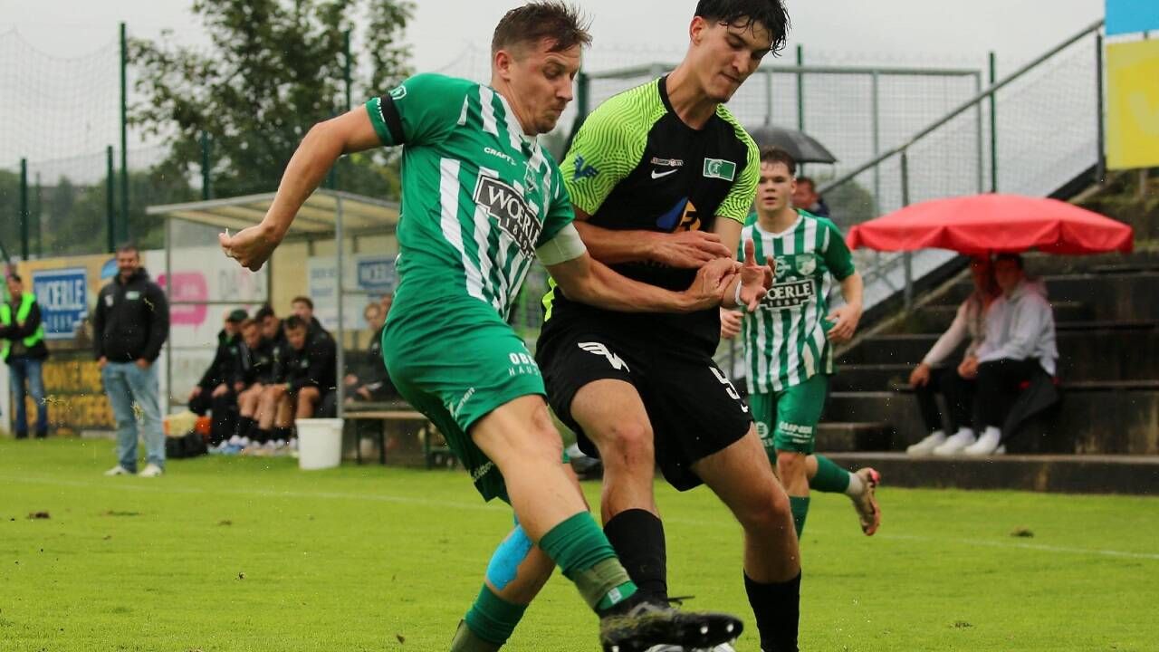 Zwei Fußballspieler sind in einem Spiel engagiert, einer trägt ein grünes Trikot und der andere ein schwarz-grünes Trikot. Beide sind in Aktion, einer versucht den Ball zu schießen, während der andere verteidigt.