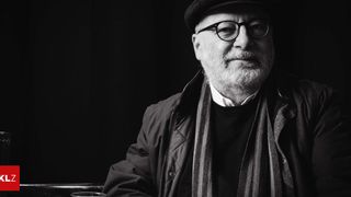 Ein schwarz-weißes Porträt eines älteren Mannes mit Brille, Mütze, Schal und Jacke, der in einem schwach beleuchteten Raum sitzt.