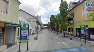 Eine leere Straße in einer Stadt mit einem Schild, das eine 20 km/h Zone anzeigt. Es gibt Bäume und Geschäfte auf beiden Seiten der Straße. Der Himmel ist bewölkt.