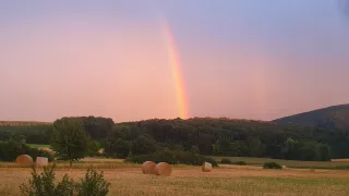 Ein farbenfroher Regenbogen erstreckt sich über einem Feld mit Heuballen und Bäumen und schafft eine ruhige und malerische Szenerie.
