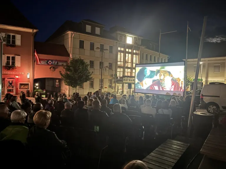 Eine Menschenmenge versammelt sich nachts im Freien, um einen Film auf einer großen Leinwand vor einem Gebäude zu sehen.