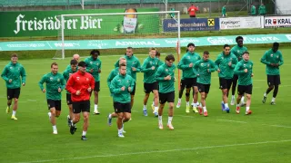 Eine Fußballmannschaft in grünen Uniformen joggt auf einem Feld, mit einem Tor und einem Zaun im Hintergrund.