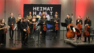 Eine Gruppe von Musikern tritt mit einem Banner auf, das Heimat Hast besagt. Die Musiker spielen Violinen, Cellos und Trompeten. Sie tragen formelle Kleidung.