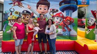 Vier Personen stehen vor einem Paw Patrol-inspirierten Aufblasbar, wobei eine Frau ein Baby und ein Mann Eintrittskarten hält.