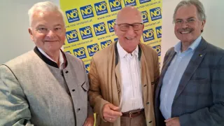 Drei Männer in formeller Kleidung stehen nebeneinander vor einem gelben Banner mit NO-Schildern und Logos.