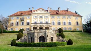 Ein großes gelbes Herrenhaus mit einem braunen Dach und mehreren Fenstern. Es steht auf einem Hügel mit gepflegten Hecken und Büschen davor.