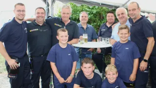 Eine Gruppe von Männern und Jungen in blauen Hemden lächelt auf einem Foto. Sie stehen unter einem Zelt. Sie posieren für ein Bild. Auf dem Tisch sind Gläser und Tassen.