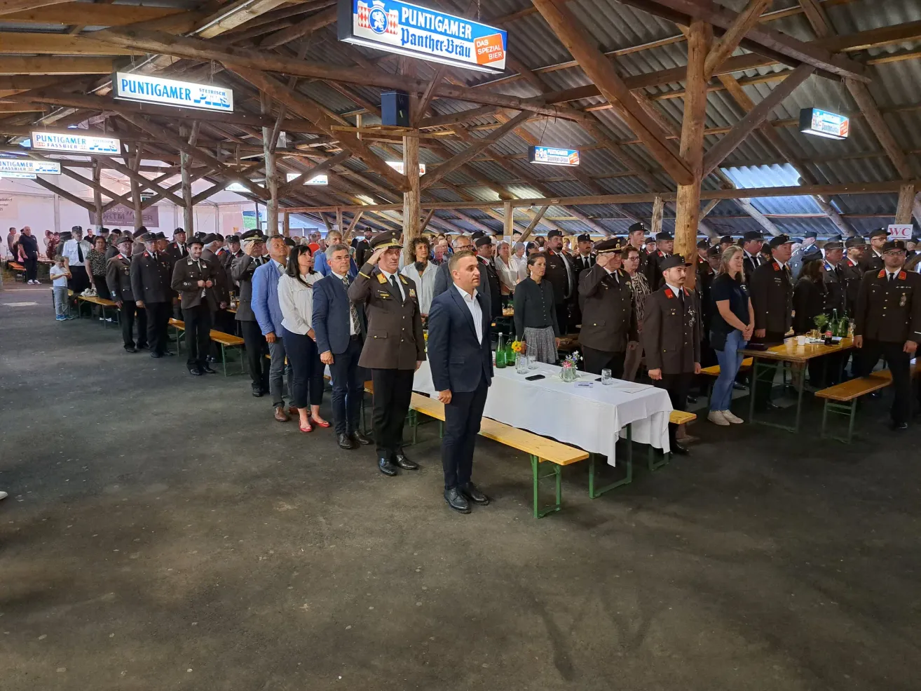 In einem großen Saal stehen viele Menschen in Uniformen und Zivilkleidung zusammen. Ein Mann in Anzug und Krawatte steht vor einem Tisch mit Blumen und Gläsern. Oben hängen Schilder von der Decke, darunter eines mit der Aufschrift 'Puntigamer'.