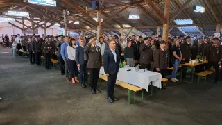 In einem großen Saal stehen viele Menschen in Uniformen und Zivilkleidung zusammen. Ein Mann in Anzug und Krawatte steht vor einem Tisch mit Blumen und Gläsern. Oben hängen Schilder von der Decke, darunter eines mit der Aufschrift 'Puntigamer'.