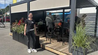Eine Frau steht vor einem überdachten Außensitzbereich. Auf beiden Seiten stehen Topfpflanzen, und eine Fahne weht im Hintergrund. Mehrere Personen sitzen an Tischen im Inneren.