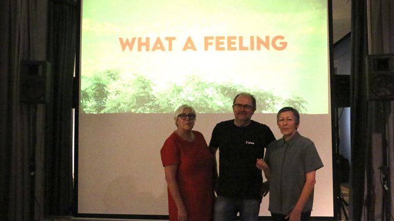 Drei Personen stehen gemeinsam vor einer Projektionsleinwand mit der Aufschrift 'What a Feeling'. Die Frau trägt ein rotes Kleid, der Mann in der Mitte ein schwarzes T-Shirt und die Frau rechts eine graue Bluse.