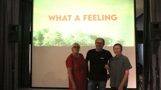 Drei Personen stehen gemeinsam vor einer Projektionsleinwand mit der Aufschrift 'What a Feeling'. Die Frau trägt ein rotes Kleid, der Mann in der Mitte ein schwarzes T-Shirt und die Frau rechts eine graue Bluse.