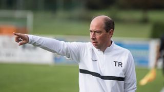 Ein Mann in einem weißen Jackett mit TR darauf gibt Anweisungen auf einem Fußballfeld.